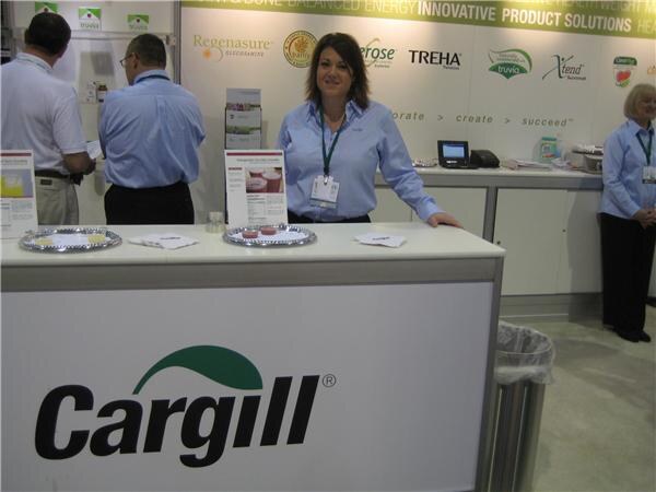 A woman in a light blue shirt with a lanyard stands behind a white Cargill counter. Signs for Regenasure, TREHA and Truvia hang behind her, with other staff at the sides.