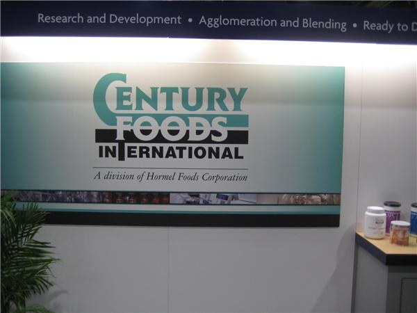 A Century Foods International sign, noting it is a division of Hormel Foods Corporation, hangs on a booth wall. Several supplement containers sit on a shelf at the right.