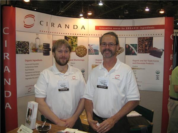 Two men in white polo shirts with exhibitor badges stand at a table in the Ciranda booth, in front of a display about organic ingredients, cocoa and chocolate products.