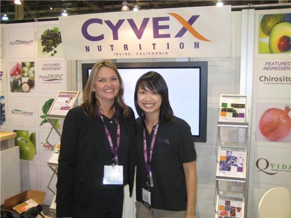 Two smiling women with lanyards stand together in the Cyvex Nutrition booth, in front of a screen, brochure racks and ingredient panels including a pomegranate.