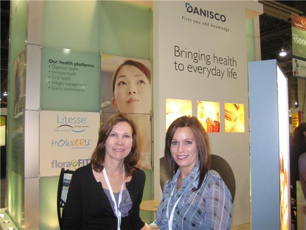 Two smiling women with lanyards stand in front of a Danisco booth reading 'Bringing health to everyday life', with Litesse, Howaru and floraFIT signs beside them.