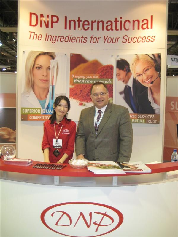 A woman in a red DNP shirt and a man in a tweed jacket and tie stand behind a red curved counter under a DNP International sign, with brochures and pens on the counter.