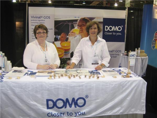 Two women in white tops with badges stand behind a table with a DOMO 'Closer to you' cloth, in front of a Vivinal GOS banner showing a person holding a glass.