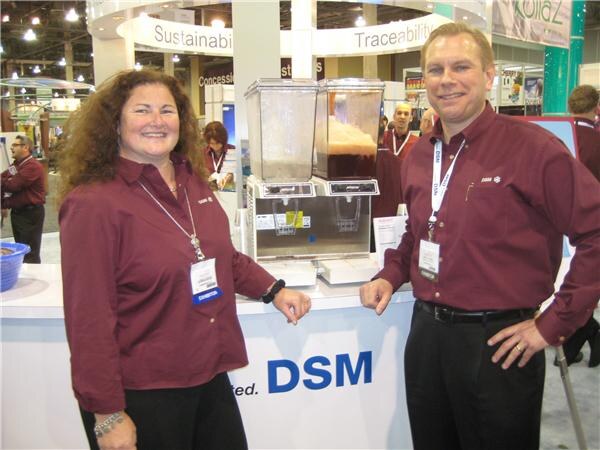 A woman and a man in maroon shirts and lanyards stand at a white DSM counter holding two beverage dispensers, with other exhibit booths behind them.