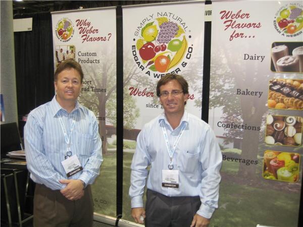 Two men in light blue dress shirts and lanyards stand in front of Edgar A. Weber & Co. banners reading "Why Weber Flavors?" and "Weber Flavors for..." with food photos.