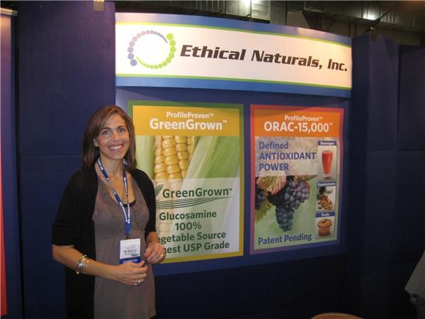 A smiling woman with a lanyard stands in front of an Ethical Naturals, Inc. booth with GreenGrown glucosamine and ORAC-15,000 panels.