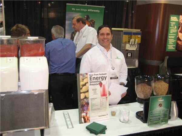 A smiling man in a white shirt stands at a Farbest counter with an Energy Smoothie Premix sign, smoothie dispensers and grain jars, while two men talk behind him.