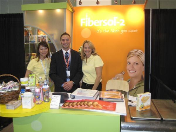 A man in a dark suit stands between two women in yellow polo shirts at a Fibersol-2 booth with an orange backdrop, with product samples and brochures on the table.