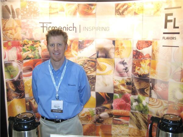 A man in a blue Firmenich shirt with an exhibitor badge stands in front of a Firmenich Flavors backdrop of food photos, with two black airpots beside him.