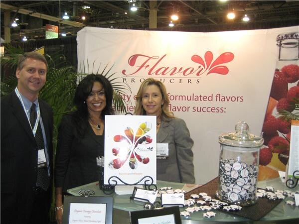 A man and two women in business attire stand at a Flavor Producers booth, with a jar of chocolate chips and organic energy chocolate signs on the table.