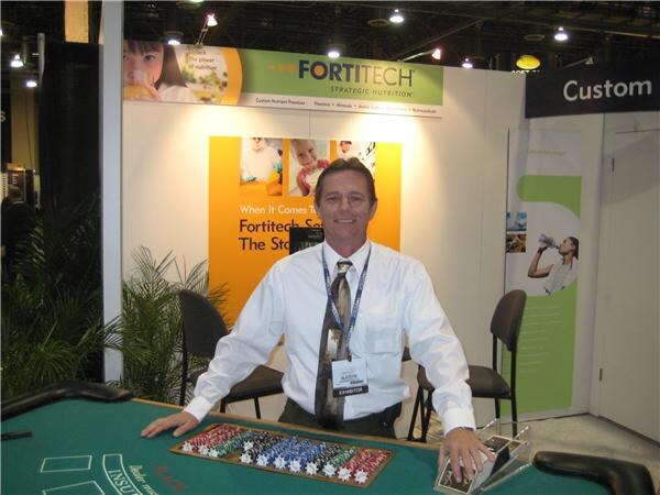 A smiling man in a white dress shirt and tie with an exhibitor badge stands behind a green blackjack table in the Fortitech Strategic Nutrition booth, with casino chips in front of him.