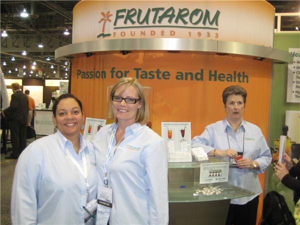 Three women in light blue shirts and lanyards stand in front of an orange Frutarom booth reading 'Passion for Taste and Health', with product cards on a glass counter.