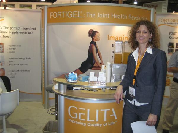 A woman with curly hair, wearing a gray blazer and a lanyard, stands beside an orange Gelita counter in a booth with Fortigel joint health banners.