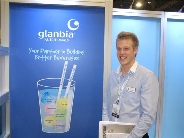 A smiling young man in a light blue shirt with an exhibitor lanyard stands before a blue Glanbia Nutritionals banner reading 'Your Partner in Building Better Beverages'.
