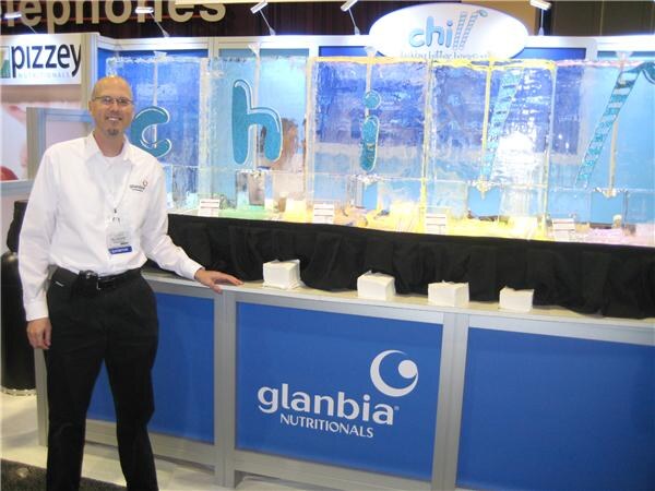 A bald man with glasses and a white shirt stands beside a Glanbia Nutritionals counter topped with ice blocks carved with the letters of 'chill'; a Pizzey Nutritionals sign is behind him.