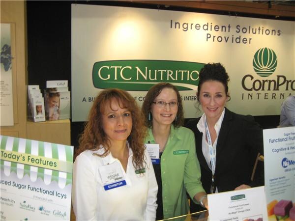 Three women stand at a counter in front of a sign reading 'Ingredient Solutions Provider' with GTC Nutrition and CornProducts International logos; signs list functional foods.