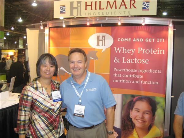 A woman in a plaid shirt and a man in a blue polo with exhibitor badges stand in front of a Hilmar Ingredients banner reading 'Whey Protein & Lactose'.