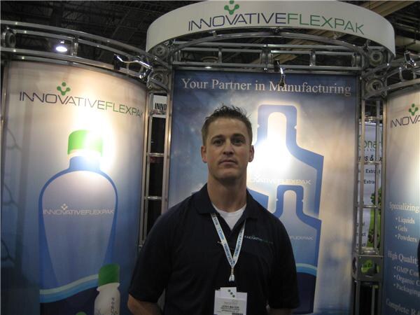 A man in a dark polo shirt with a lanyard stands in front of Innovative Flexpak banners showing bottle graphics and the line 'Your Partner in Manufacturing'.