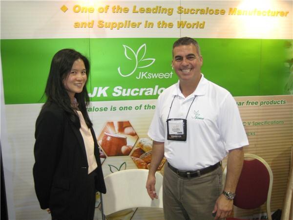 A woman in a black jacket and a smiling man in a white polo stand in front of a green JKsweet banner that reads 'One of the Leading Sucralose Manufacturer and Supplier in the World'.