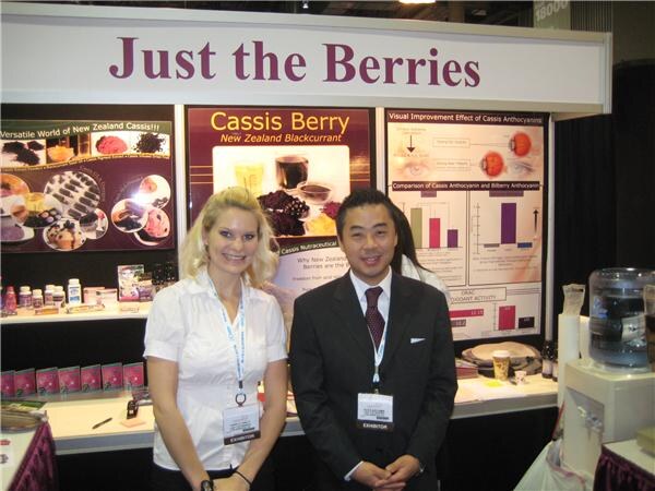 A blonde woman and a man in a dark suit, both wearing exhibitor badges, stand in the Just the Berries booth in front of posters about cassis berry and cassis anthocyanin.