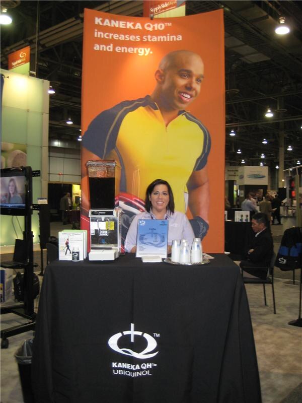 A woman sits behind a black draped table marked Kaneka QH Ubiquinol, in front of an orange banner reading Kaneka Q10 increases stamina and energy, with a smiling man in a yellow shirt.