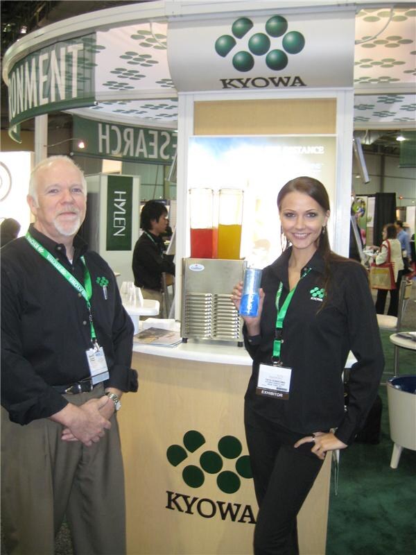 A gray-haired man with a beard and a woman holding a blue can stand at a round Kyowa booth counter with two juice dispensers. Both wear green lanyards and exhibitor badges.