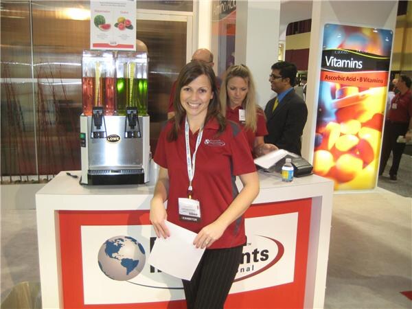A smiling woman in a red polo and exhibitor badge stands at a booth counter with a Lowe dispenser holding red and green drinks; two people and a Vitamins banner are behind her.