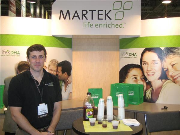 A man in a black polo with an exhibitor badge stands in the Martek booth, near a table with a bottle of juice, stacked cups and small samples, with life'sDHA banners behind him.