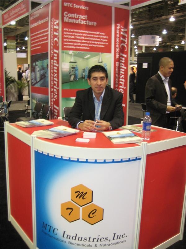 A man in a dark jacket sits at a red counter in the MTC Industries, Inc. booth, with brochures and a water bottle in front of him. Another man in a suit stands behind him.