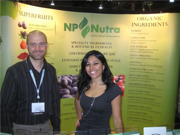 A bald bearded man and a smiling dark-haired woman stand in the NP Nutra booth in front of a display listing superfruits and organic ingredients. The man wears an exhibitor badge.