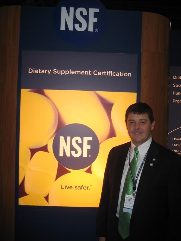 A man in a dark suit and green tie with a badge stands beside a large NSF display reading Dietary Supplement Certification and Live safer, with yellow tablets pictured.
