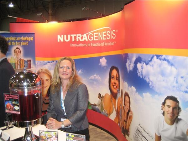 A blonde woman in a gray jacket stands in the Nutragenesis booth next to a large metal beverage dispenser, with banners showing smiling people against a blue sky.