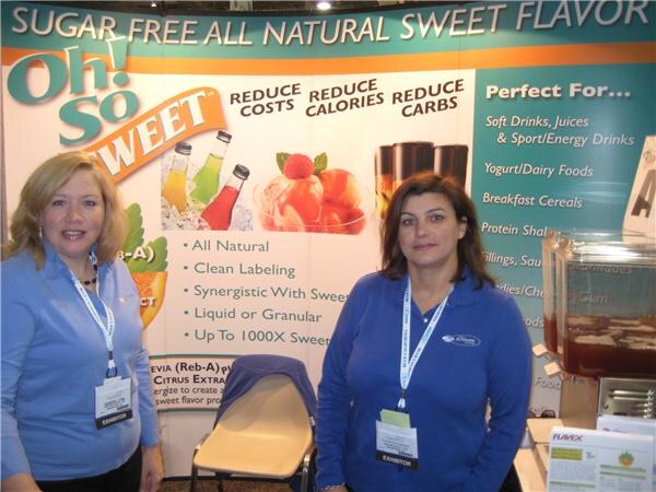Two women in blue shirts with exhibitor badges stand in front of an Oh! So Sweet banner reading Sugar Free All Natural Sweet Flavor. Flavex literature sits on a table at right.