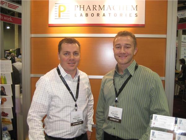 Two smiling men wearing lanyards and exhibitor badges stand side by side in front of a wood-paneled Pharmachem Laboratories sign.