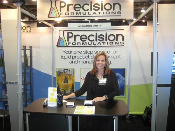 A woman with shoulder-length brown hair and an exhibitor badge stands behind a table in the Precision Formulations booth, under signs reading Precision Formulations.