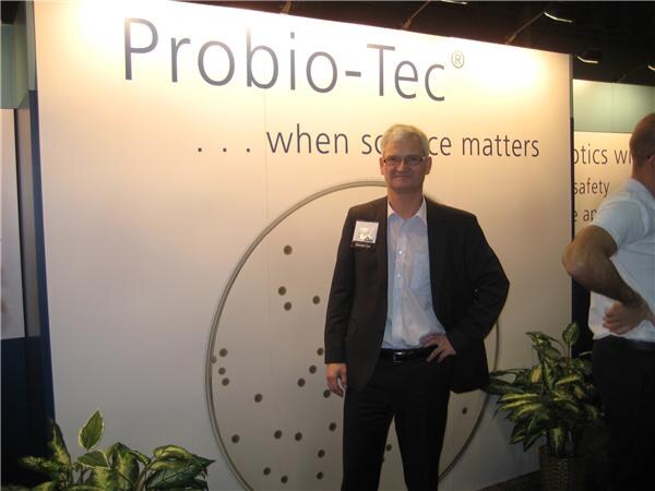 A man in glasses and a dark blazer stands with a hand on his hip in front of a Probio-Tec wall reading when science matters. Part of another person is visible at the right.