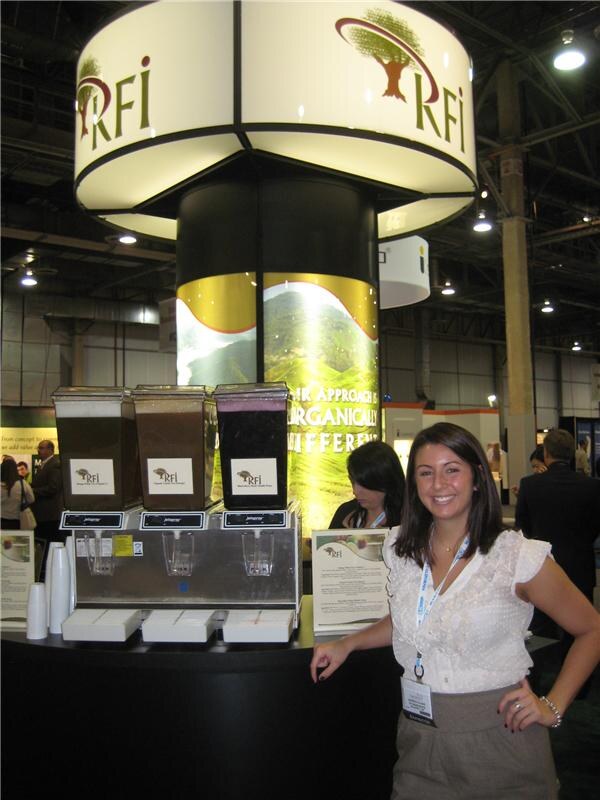 A smiling woman with dark hair in a white blouse and lanyard stands beside three beverage dispensers with RFI labels under a large RFI sign with a tree logo.