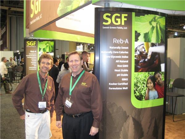 Two men in brown SGF shirts and green lanyards stand beside a tall SGF Sweet Green Fields sign listing Reb-A features, with other attendees in the background.