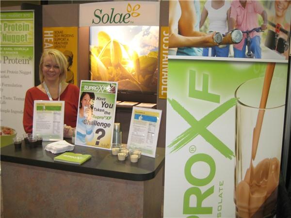 A blonde woman in a red top stands behind a counter at the Solae booth, with Supro XF signs, a Supro XF Challenge poster and small sample cups on the counter.