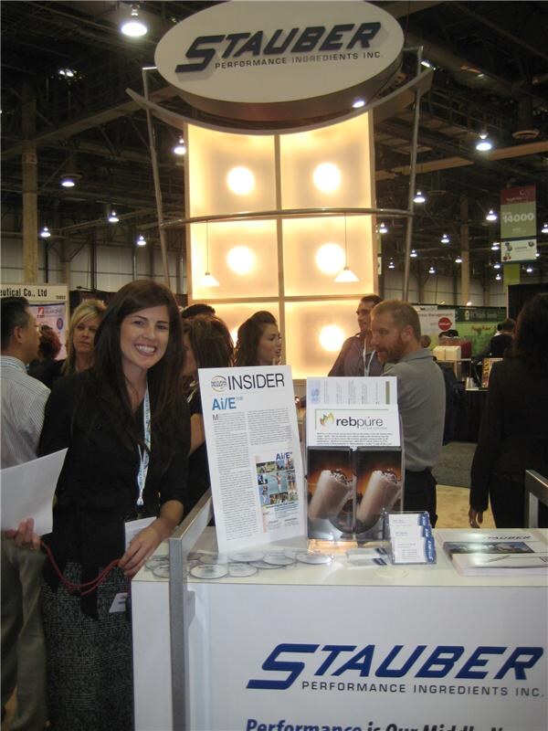 A smiling woman with long dark hair stands at the Stauber Performance Ingredients booth counter, which displays rebpüre literature and discs; other attendees stand behind her.