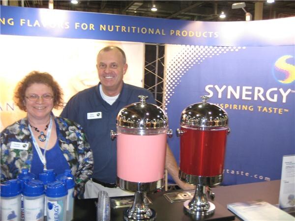 A woman with curly hair and glasses and a man in a blue shirt stand behind a counter with two drink dispensers, in front of a Synergy Inspiring Taste banner.