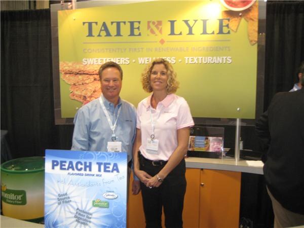 A man in a blue shirt and a woman in a pink polo stand in front of a Tate & Lyle sign, next to a Peach Tea drink mix display and a Promitor container.