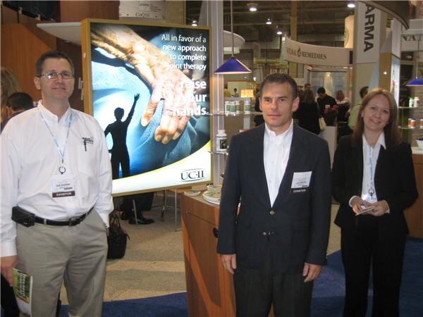 Two men and a woman in business attire with exhibitor badges stand beside a lit UC-II display showing an elderly hand and the words 'raise your hands'.