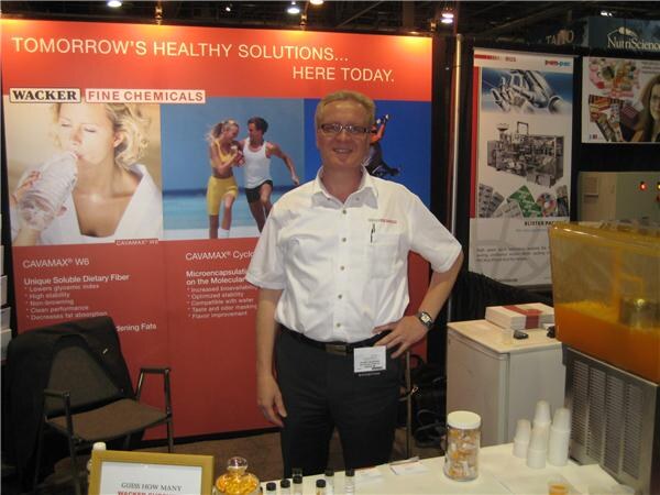 A man with glasses in a white shirt stands with hands on hips in the Wacker Fine Chemicals booth, in front of a Tomorrow's Healthy Solutions banner.