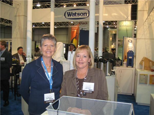 Two women with exhibitor badges stand together in front of a Watson sign in a trade show booth, with other attendees and booth equipment behind them.