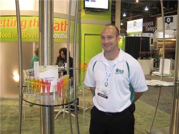 A smiling man in a white and green Wild polo and exhibitor badge stands beside a glass table holding colorful drink samples in small tubes, with other booths behind him.
