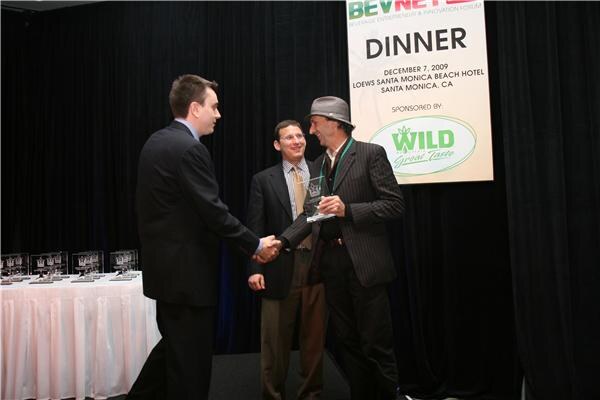 A man in a suit shakes hands with a man in a gray hat holding a glass award, while a third man looks on, beneath a dinner banner sponsored by Wild Great Taste.