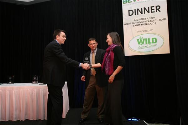 A man in a dark suit shakes hands with a woman in a magenta scarf as a man in a tie stands between them holding an award, beneath a Wild Great Taste dinner banner.