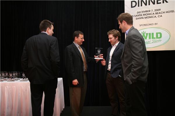 Four men in dark jackets stand on stage as one hands a glass award to another, with a table of awards at left and a dinner banner sponsored by Wild above.
