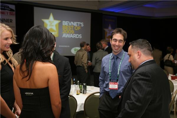 Guests talk at the reception in front of a screen reading BevNET's Best of 2009 Awards. A woman in the foreground wears a black Monster Energy top, and a man wears a badge.